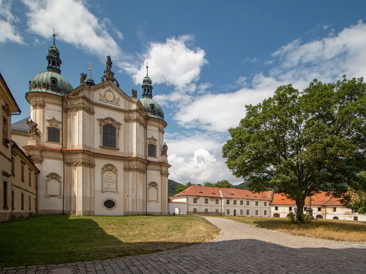 Barokní kostel se dvěma zelenými kupolemi a sochami na průčelí, před ním travnaté nádvoří s dlážděnou cestou a vzrostlým stromem; vpravo dlouhá budova s červenou střechou a zalesněný kopec v pozadí